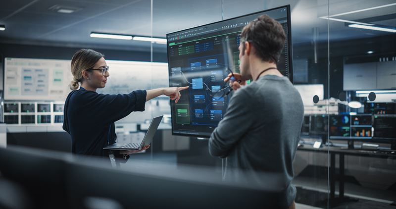 A woman in a dark shirt points at a computer screen while a man in a grey shirt looks at the screen.